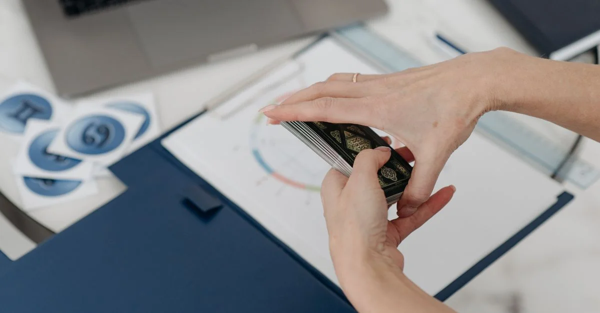 Una mujer barajando cartas de tarot sobre un escritorio con un gráfico del horóscopo y una laptop.