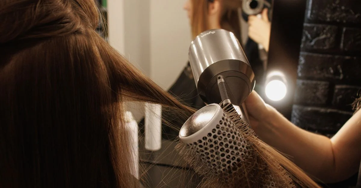 Une coiffeuse sèche les cheveux d'une femme avec une brosse et un sèche-cheveux dans un salon.