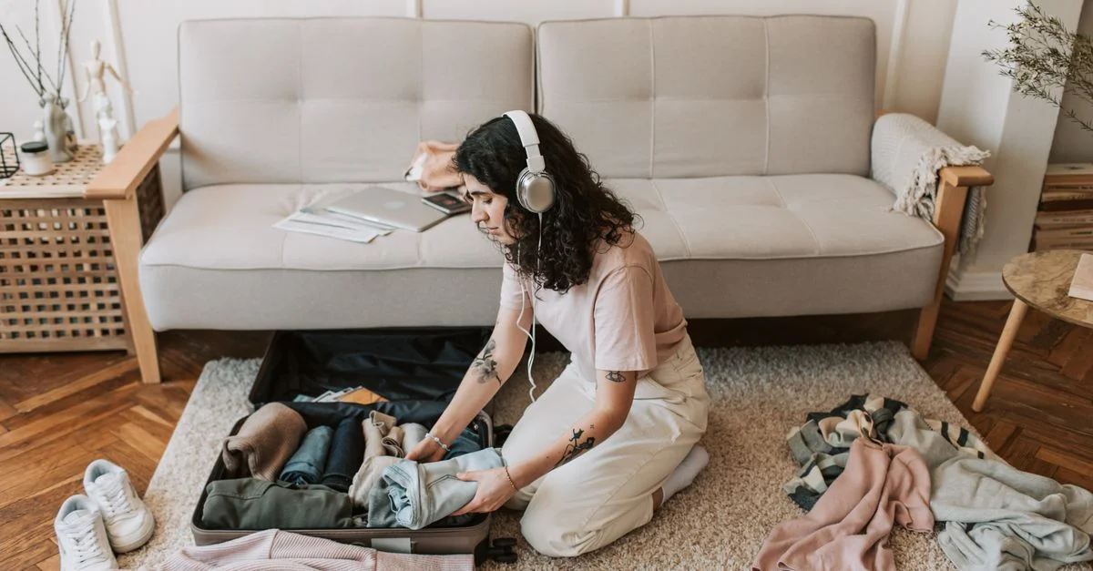Mujer empacando una maleta en el suelo de la sala, usando auriculares, preparándose para un viaje.