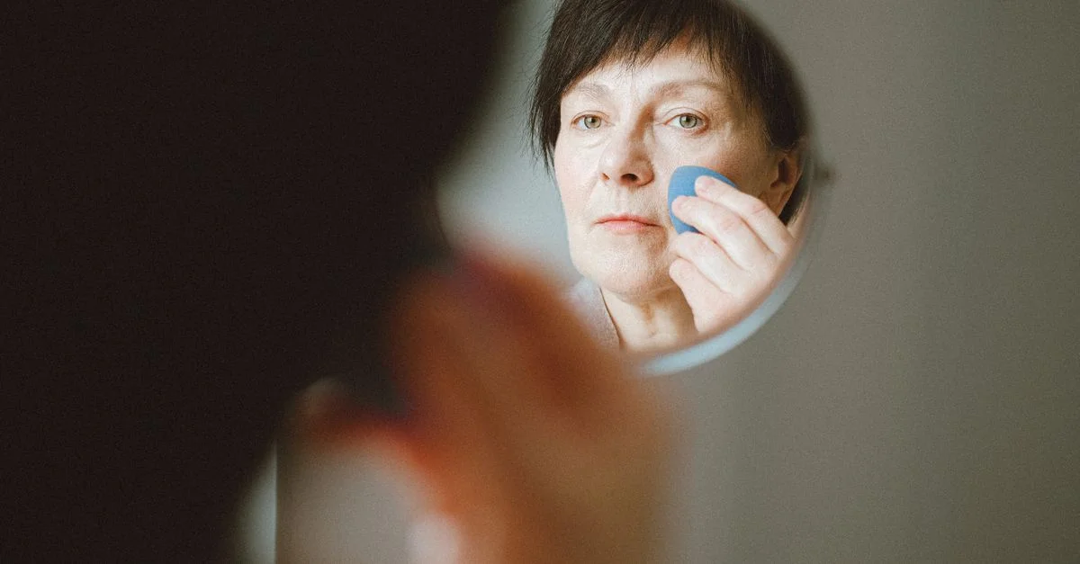 Mujer anciana mirándose en el espejo mientras aplica una esponja de maquillaje.