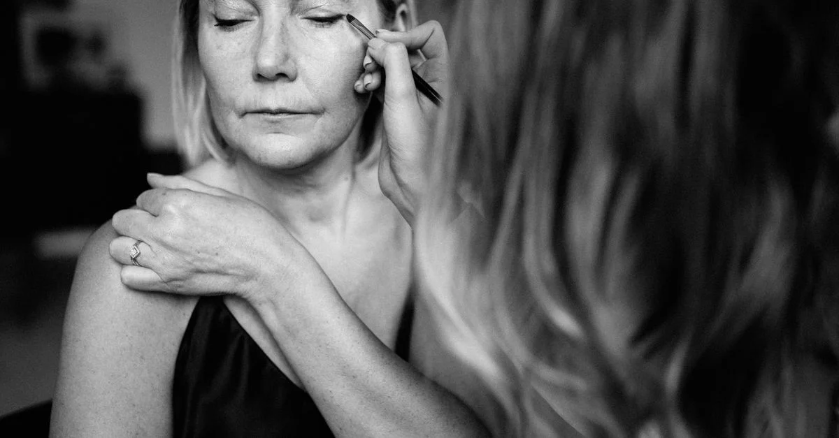 Una mujer con los ojos cerrados, recibiendo la aplicación de maquillaje en una foto en blanco y negro.